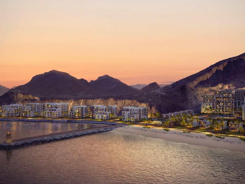 Stadt am Meer mit Bergen im Hintergrund, beleuchtete Gebäude bei Sonnenuntergang, ruhiges Wasser.