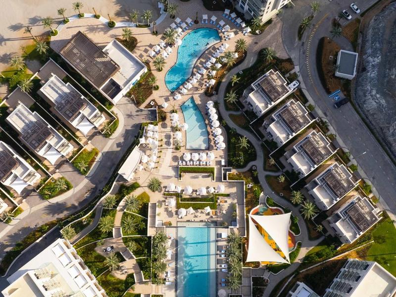 Luftaufnahme einer Hotelanlage mit mehreren Pools, Sonnenschirmen und angrenzenden Gebäuden am Strand.
