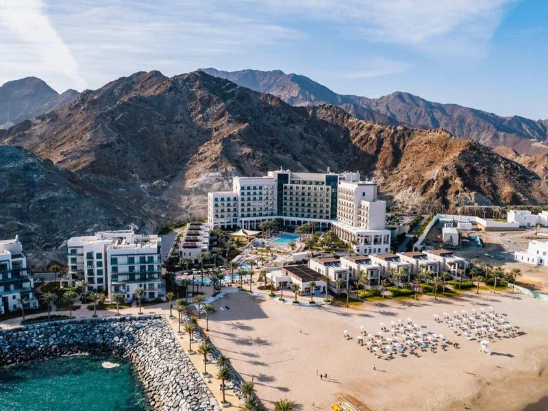 Luxushotel am Strand mit Liegestühlen und Bergen im Hinterland unter blauem Himmel.