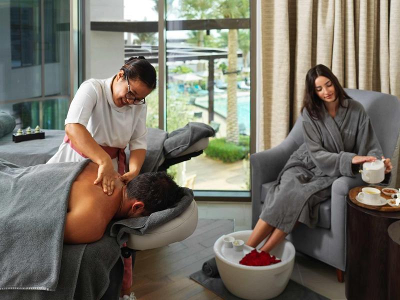 Ein Mann massiert einen Rücken, während eine Frau daneben in Bademantel sitzt und Füße im Becken badet.