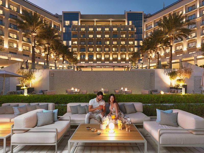 Paar sitzt abends auf gemütlicher Terrasse vor beleuchtetem großen Hotel mit Palmen und eleganten Sitzmöbeln.