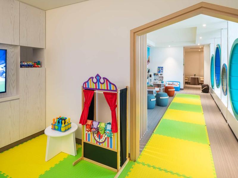 Kinderspielzimmer mit buntem Schaumstoffboden, Spielküche, Fernsehbildschirm und runden Fenstern.