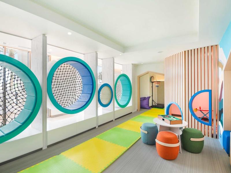 Moderner, bunter Raum mit runden Fenstern, grünen Sitzhockern und gepolstertem Boden.