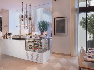 Modernes Café mit weißem Tresen, Glastheke, Pendelleuchten und großen Fenstern mit Pflanzen draußen.