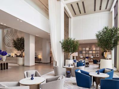 Moderne Hotellobby mit weißen Wänden, blauen Sesseln, Pflanzen und hellen Tischen in großzügigem Raum.