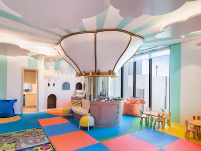 Kinderspielzimmer mit buntem Boden, Sitzkissen, Holztischen, Burgwand und großem Glühbirnen-Lüfter.