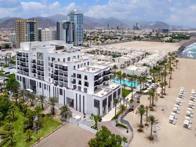 Moderner Hotelkomplex mit Pool, Palmen und Liegestühlen an einem sandigen Strand bei sonnigem Himmel.