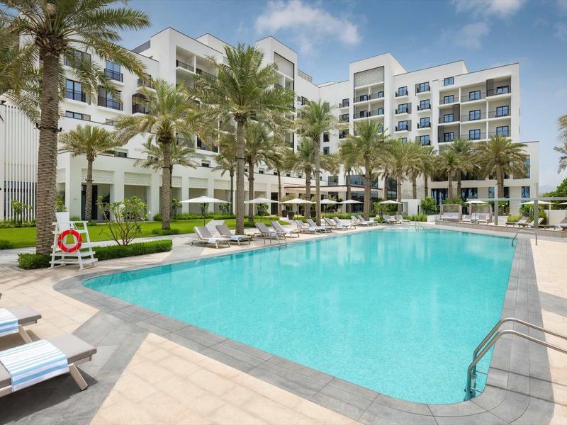 Moderner Hotelpool mit klar blauem Wasser, Palmen, Liegen und weißem Gebäude im Hintergrund.