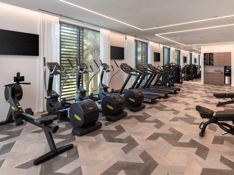 Moderner Fitnessraum mit Fitnessgeräten, hellen Wänden, großen Fenstern und gemustertem Boden.