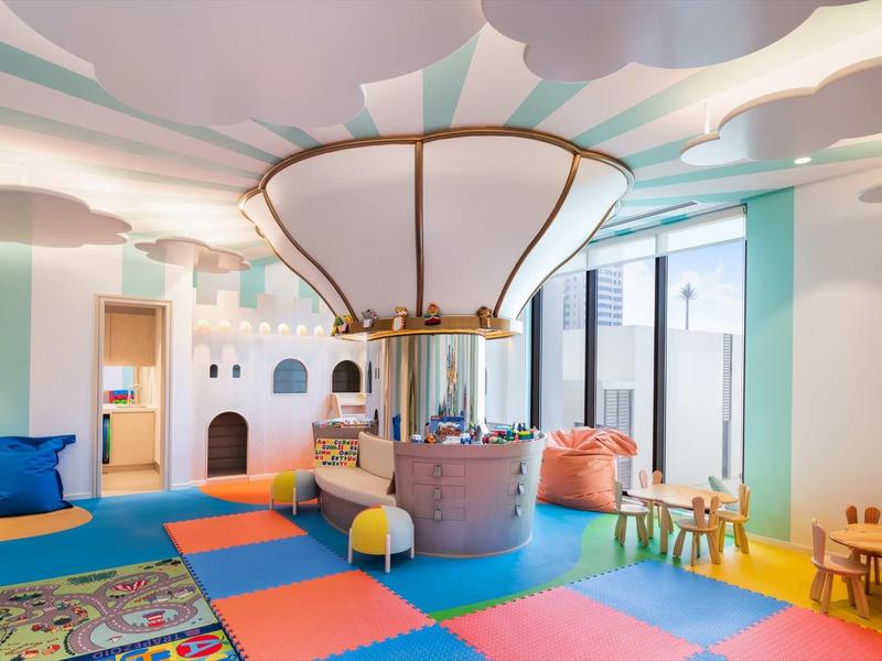 Kinderspielzimmer mit buntem Boden, Sitzkissen, Holztischen, Burgwand und großem Glühbirnen-Lüfter.