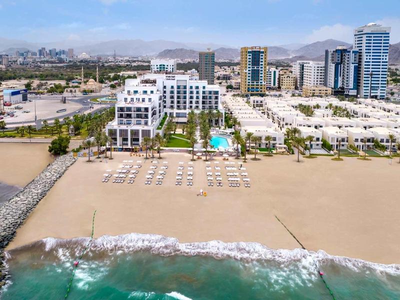 Breiter Sandstrand mit Liegestühlen vor modernen weißen Gebäuden und hohen Wolkenkratzern im Hintergrund.