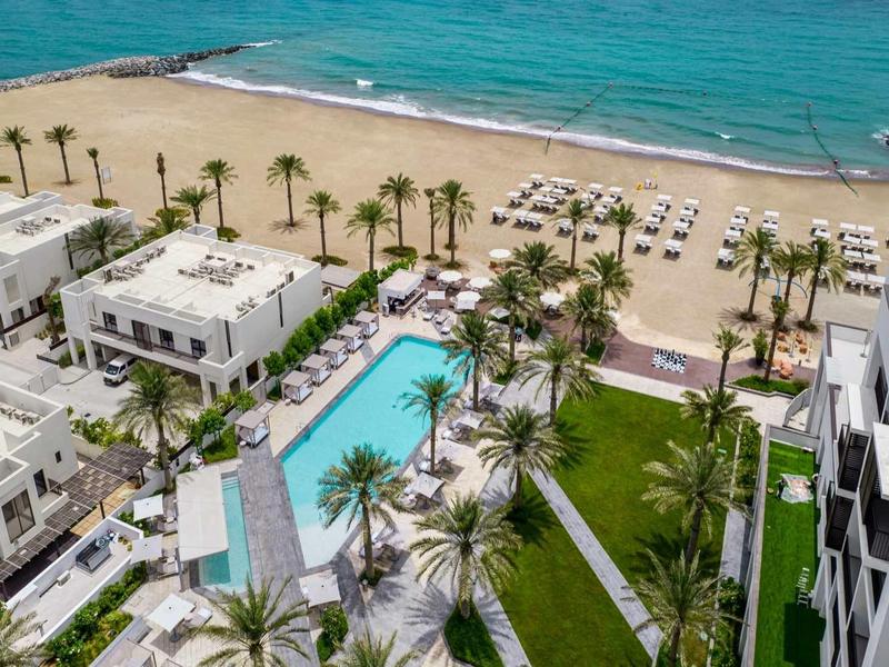 Hotel mit Pool, Palmen, Liegestühlen am Sandstrand und türkisblauem Meer im Hintergrund.