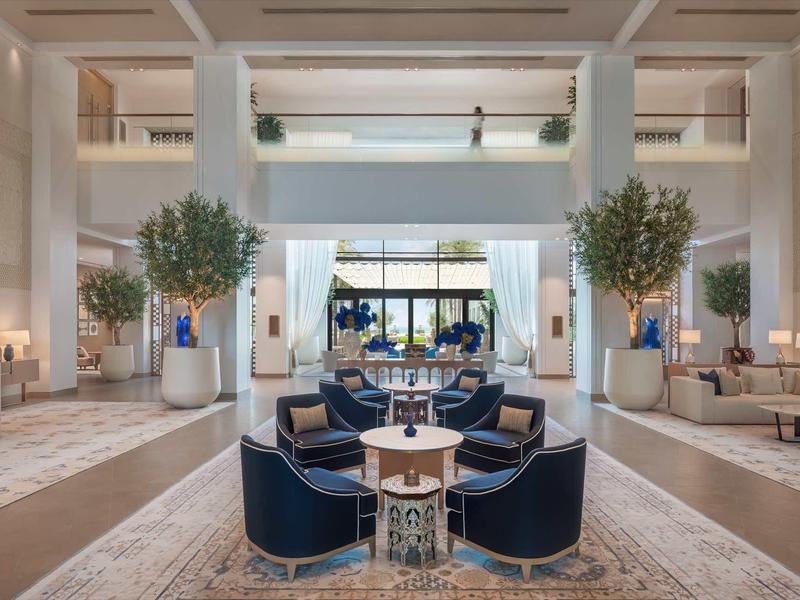 Großer, heller Hotel-Lobbybereich mit blauen Sesseln, Bäumen und moderner Beleuchtung.