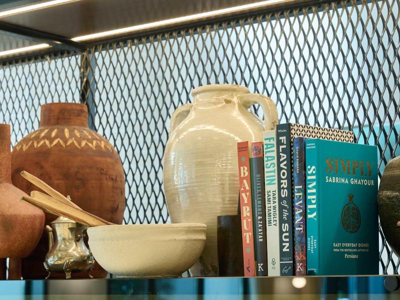 Regal mit Tontöpfen, Schüssel, Holzlöffeln und mehreren bunten Büchern vor Gittern.