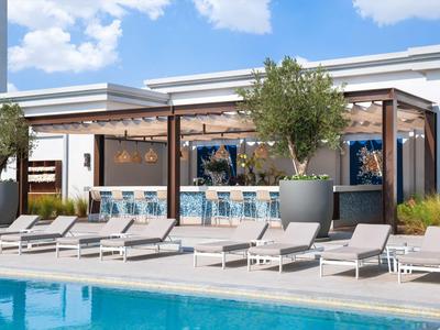 Moderne Außenbar mit Sitzplätzen und Liegestühlen neben einem Pool unter blauem Himmel.