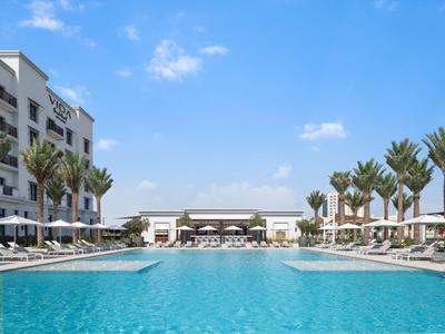 Großer blauer Pool mit weißen Liegen, Palmen und mehrstöckigem Gebäude unter blauem Himmel.