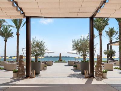 Blick von einer schattigen Terrasse mit Sitzgelegenheiten auf einen Pool und dahinter das Meer mit Palmen