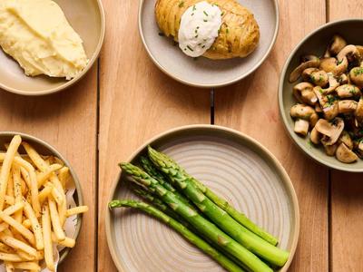 Verschiedene Speisen wie Spargel, Pommes, Kartoffelpüree, Spinat, Brötchen und Pilze auf Holzoberfläche.