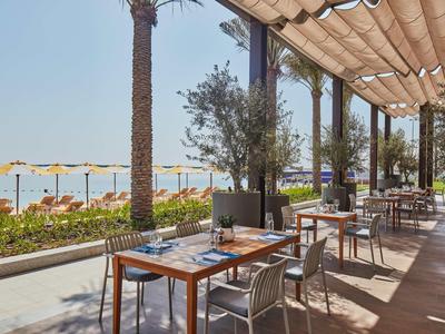 Terrassenrestaurant mit Holztischen, Metallstühlen, Palmen und Ausblick auf Strand mit Sonnenschirmen.
