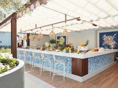 Moderner Außenbarbereich mit blauen Fliesen, weißen Barhockern und Pflanzendekoration unter Sonnendach.