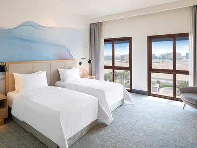 Helles Hotelzimmer mit zwei Einzelbetten, großen Fenstern, grauem Teppich und schlichtem Mobiliar.