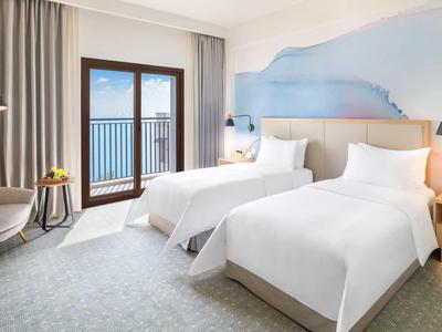 Helles Hotelzimmer mit zwei Einzelbetten, Balkon, grauem Teppich und pastellfarbenen Wänden.