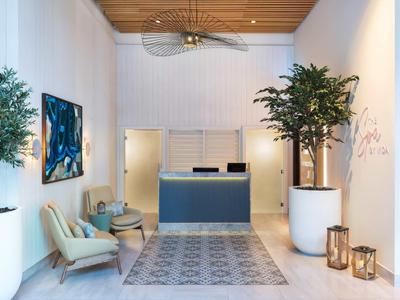 Heller Empfangsraum mit blauem Tresen, zwei Sesseln, Pflanzen und modernem Teppich auf weißem Boden.