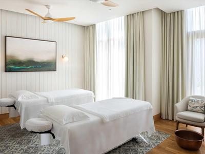 Heller, moderner Massageraum mit zwei weißen Liegen, Teppich, Stuhl, Vorhängen und großem Fenster.