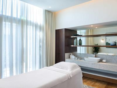 Heller Massageraum mit weißen Betten, großen Fenstern und modernen Regalen mit Dekoration.