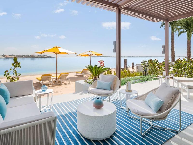 Moderne Terrasse mit weißen Möbeln, blauem Teppich, Blick auf Wasser und gelbem Sonnenschirm.