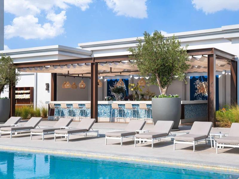 Moderne Poollandschaft mit Liegestühlen, Bar und großen Pflanzkübeln unter blauem Himmel.