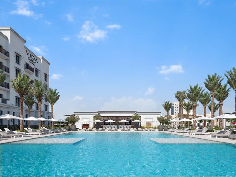 Großer blauer Pool mit weißen Liegen, Palmen und mehrstöckigem Gebäude unter blauem Himmel.