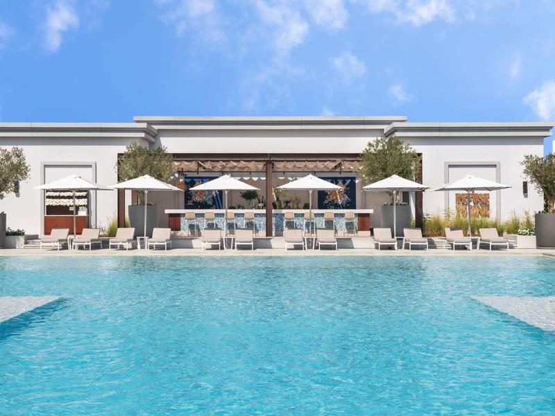 Großer Pool vor einem weißen Gebäude mit Sonnenschirmen und Liegestühlen unter blauem Himmel.