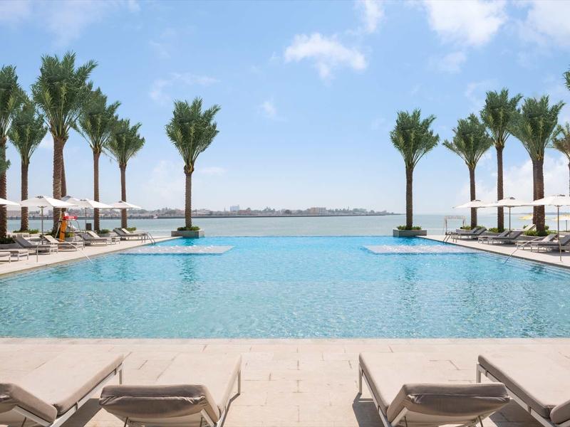 Großer Pool mit blauer Wasserfläche, umgeben von Palmen und Liegestühlen unter blauem Himmel.