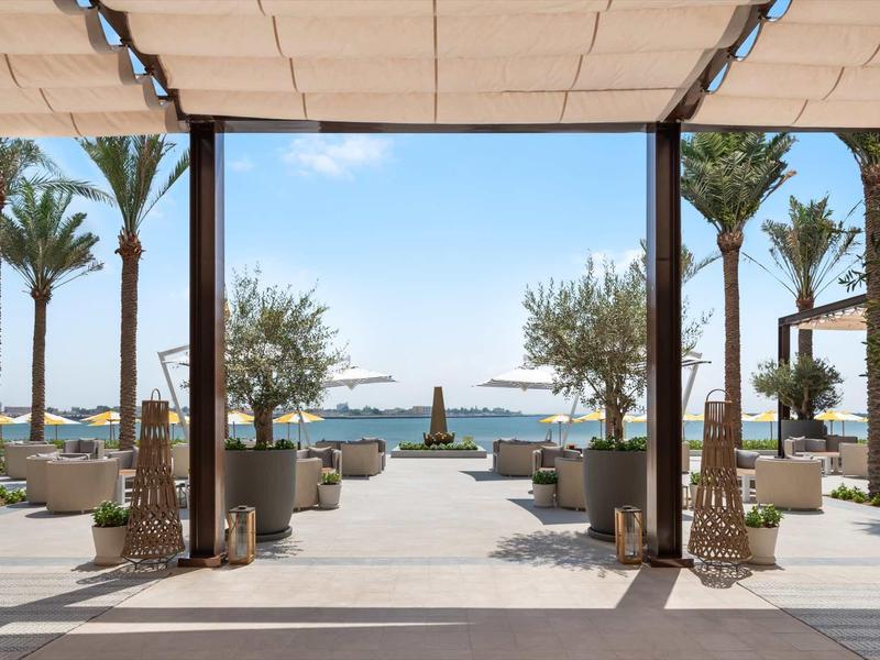 Moderner Außenbereich mit Palmen, Sitzbereichen und Blick auf das Meer unter blauem Himmel.