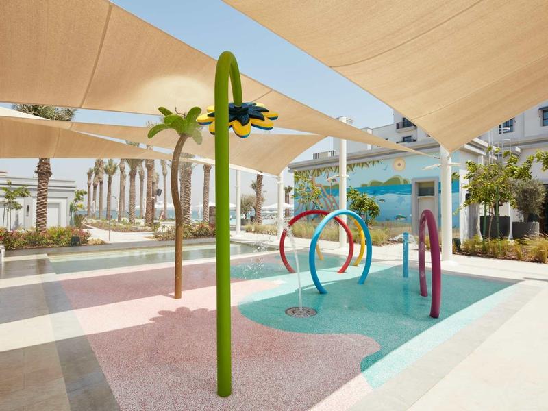 Moderner Wasserspielplatz mit bunten Sprühvorrichtungen, Sonnensegeln und Palmen im Hintergrund.