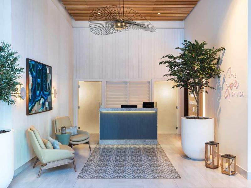 Heller Empfangsraum mit blauem Tresen, zwei Sesseln, Pflanzen und modernem Teppich auf weißem Boden.