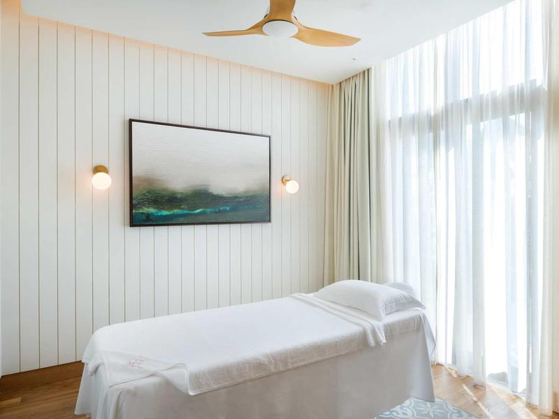 Helles Behandlungszimmer mit Massageliege, großen Fenstern, weißen Wänden und Deckenventilator.