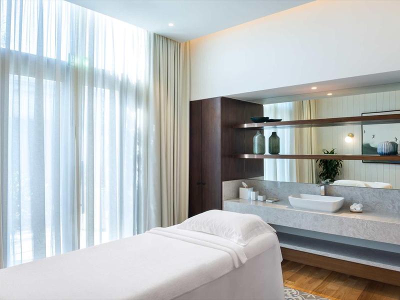 Heller Massageraum mit weißen Betten, großen Fenstern und modernen Regalen mit Dekoration.