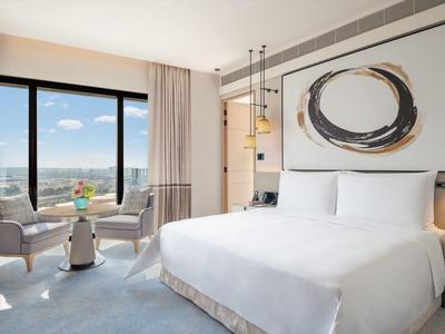 Modern hotel room with large bed, artwork, and seating area by window with city view.