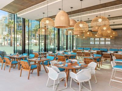 Modern hotel restaurant with large windows, wooden accents, and woven lamps above blue chairs.