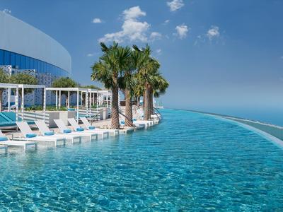 Piscine à débordement avec transats et palmiers à côté d'un hôtel moderne avec vue sur la mer.