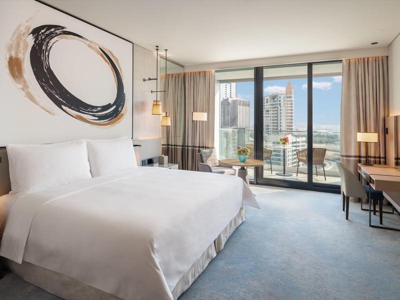 Bright hotel room with large bed, modern artwork, and city view through glass windows.