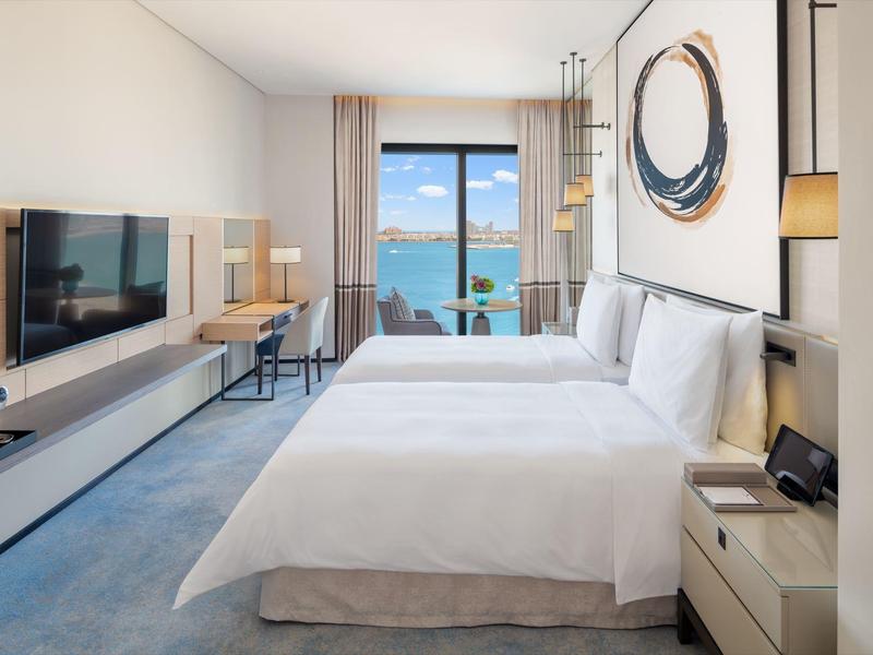 Modern hotel room with two beds, balcony, and sea view in daylight.