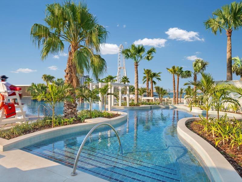 A clear pool with palm trees and a lifeguard under a blue sky in a tropical resort.