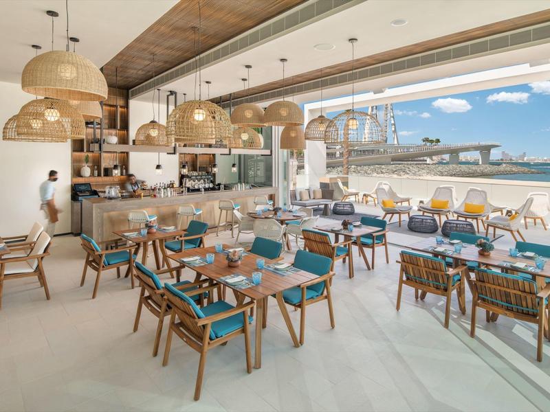 Bright, modern hotel bar with wooden chairs, blue cushions, and large windows overlooking the sea.