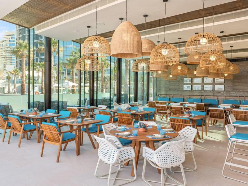 Modern hotel restaurant with large windows, wooden accents, and woven lamps above blue chairs.