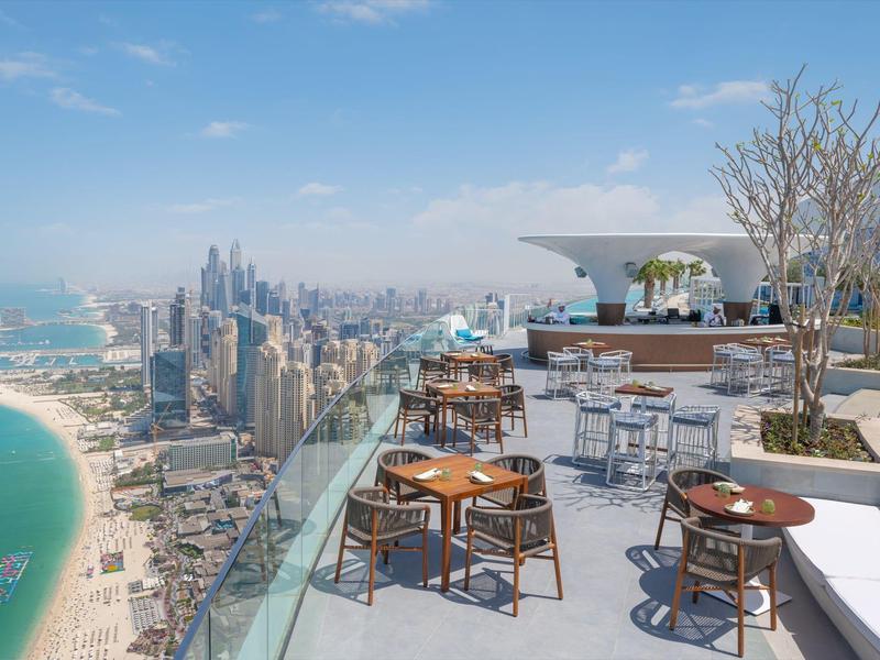 Rooftop terrace with tables, chairs, and bar overlooking beach and skyscrapers under clear sky