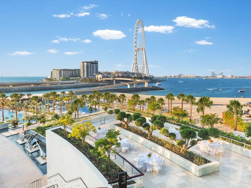 View of a promenade with palm trees, sandy beach, and Ferris wheel by the sea under blue sky