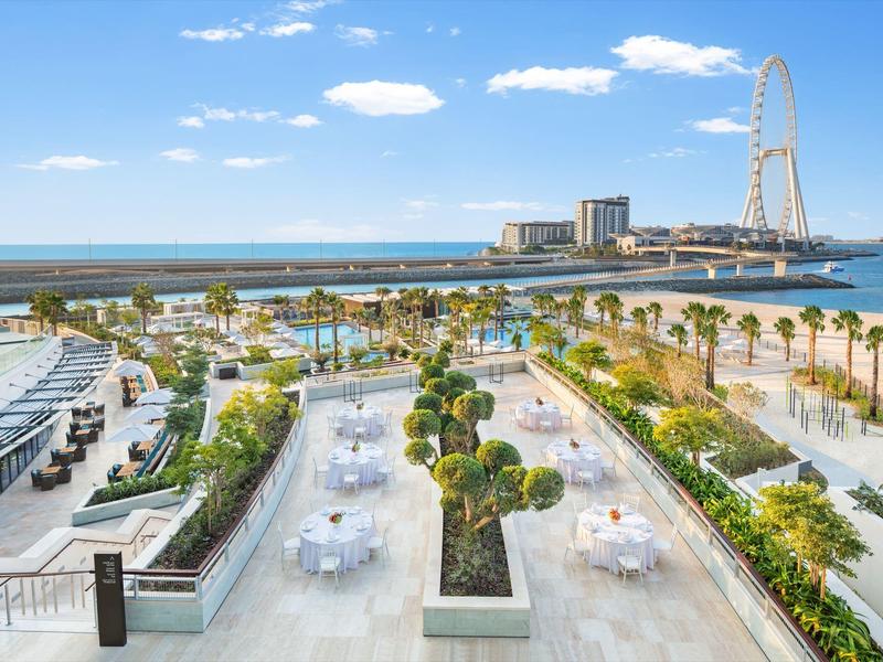 View of terrace with tables, palm trees and sea in the background with Ferris wheel under clear sky